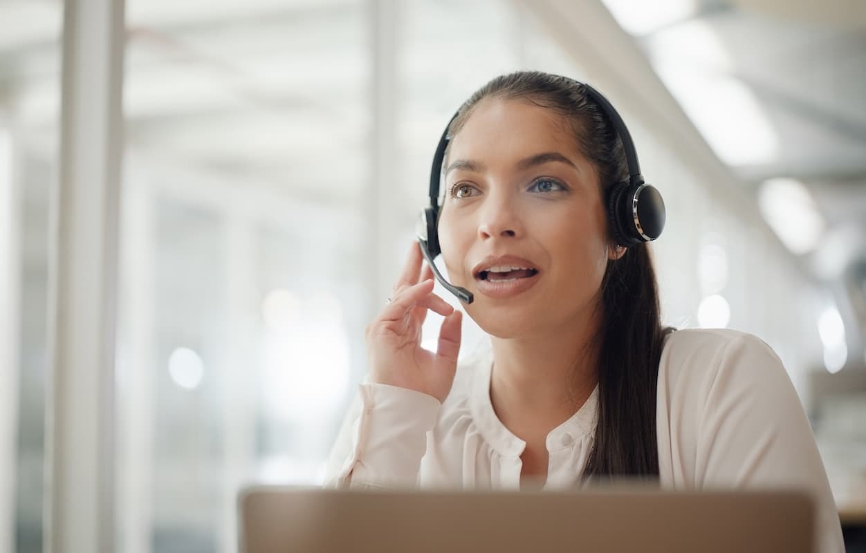 A woman wearing a black headset speaking into the microphone while looking at a laptop in a bright, modern office environment.
