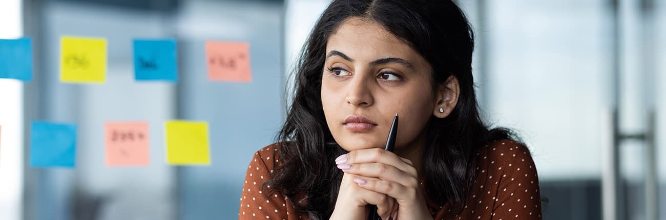 A woman with long dark hair is seated indoors, wearing a brown top with white polka dots. Her hands are clasped together near her chin. In the background, there is a glass wall with colorful sticky notes in yellow, blue, and orange arranged in rows. The setting is an office or meeting space with a modern design.