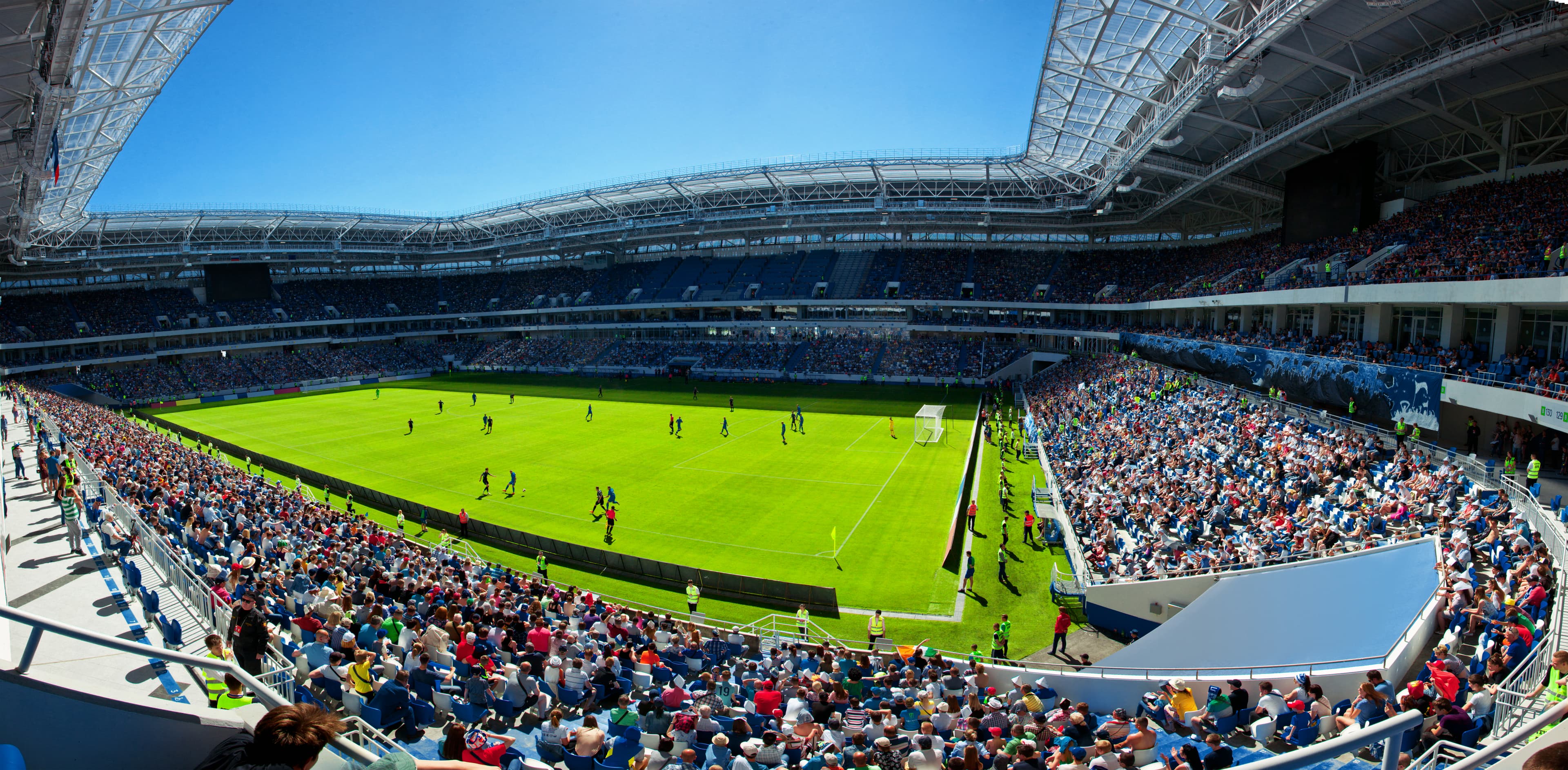 Estádio de futebol lotado durante uma partida, com arquibancadas cheias e jogadores em campo sob céu azul.