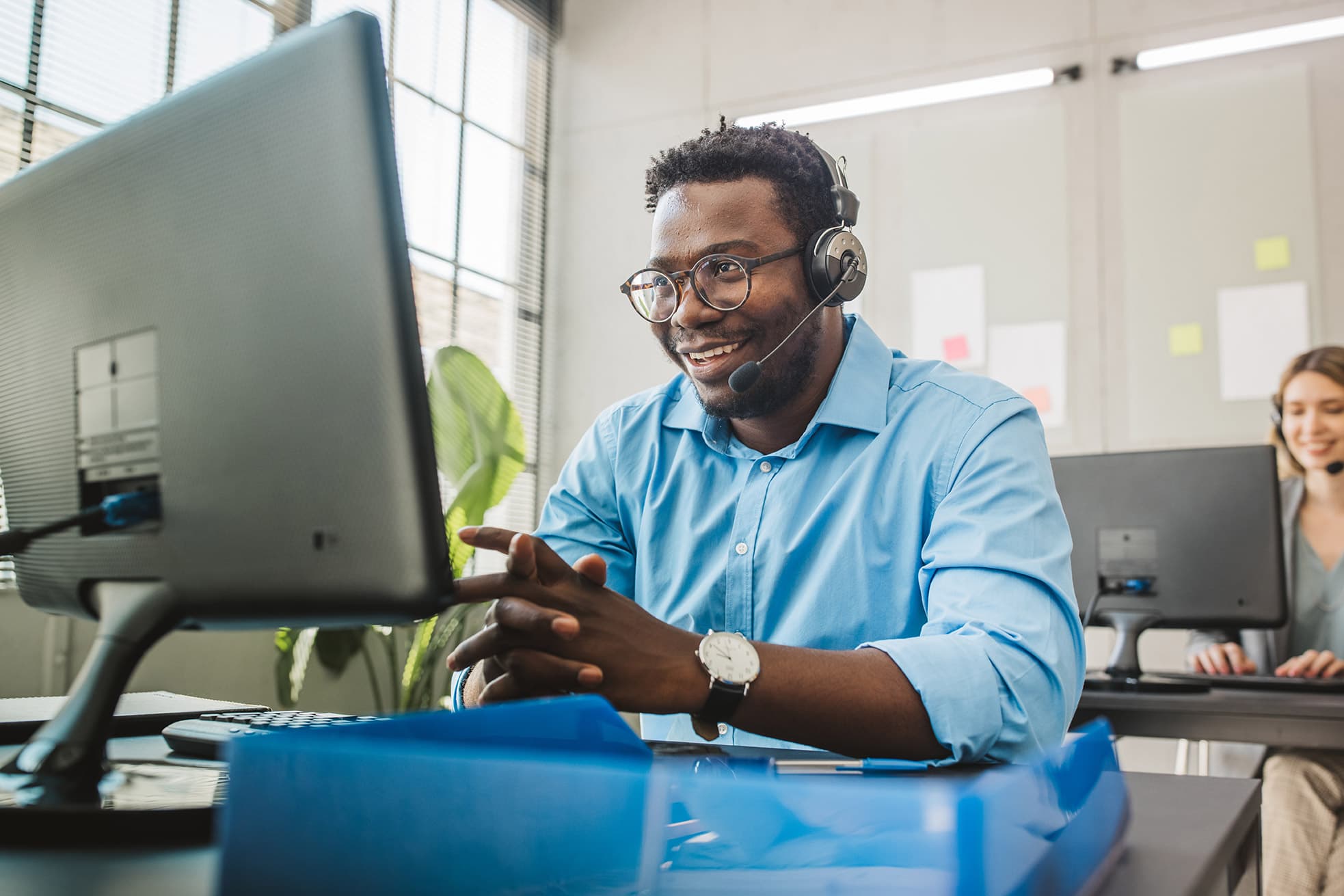 A man wearing a telephone headset and a blue button-down shirt sits at a desk, hands clasped, looking at a large computer monitor in a modern office. Other people work at computers in the background, with bright lighting and glass walls in a professional workplace.