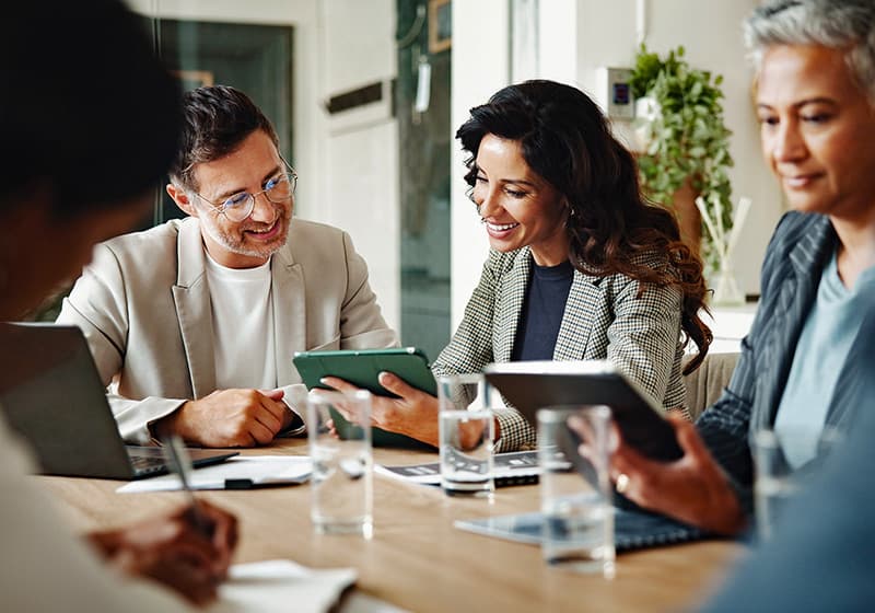 Five colleagues sit around a conference table in a modern office, reviewing information on tablets and laptops during a collaborative meeting, with notebooks and glasses of water on the table.