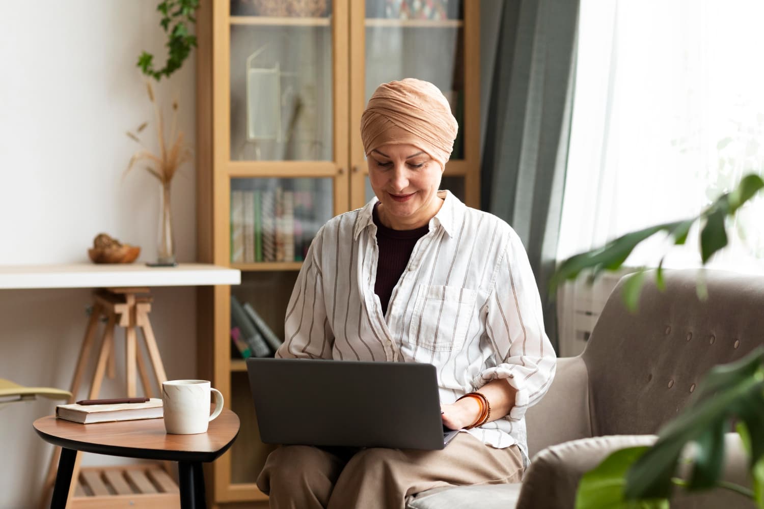 Mulher com um lenço em sua cabeça, utilizando um notebook em casa, em ambiente iluminado e confortável, representando cuidado com a saúde e acesso à informação.