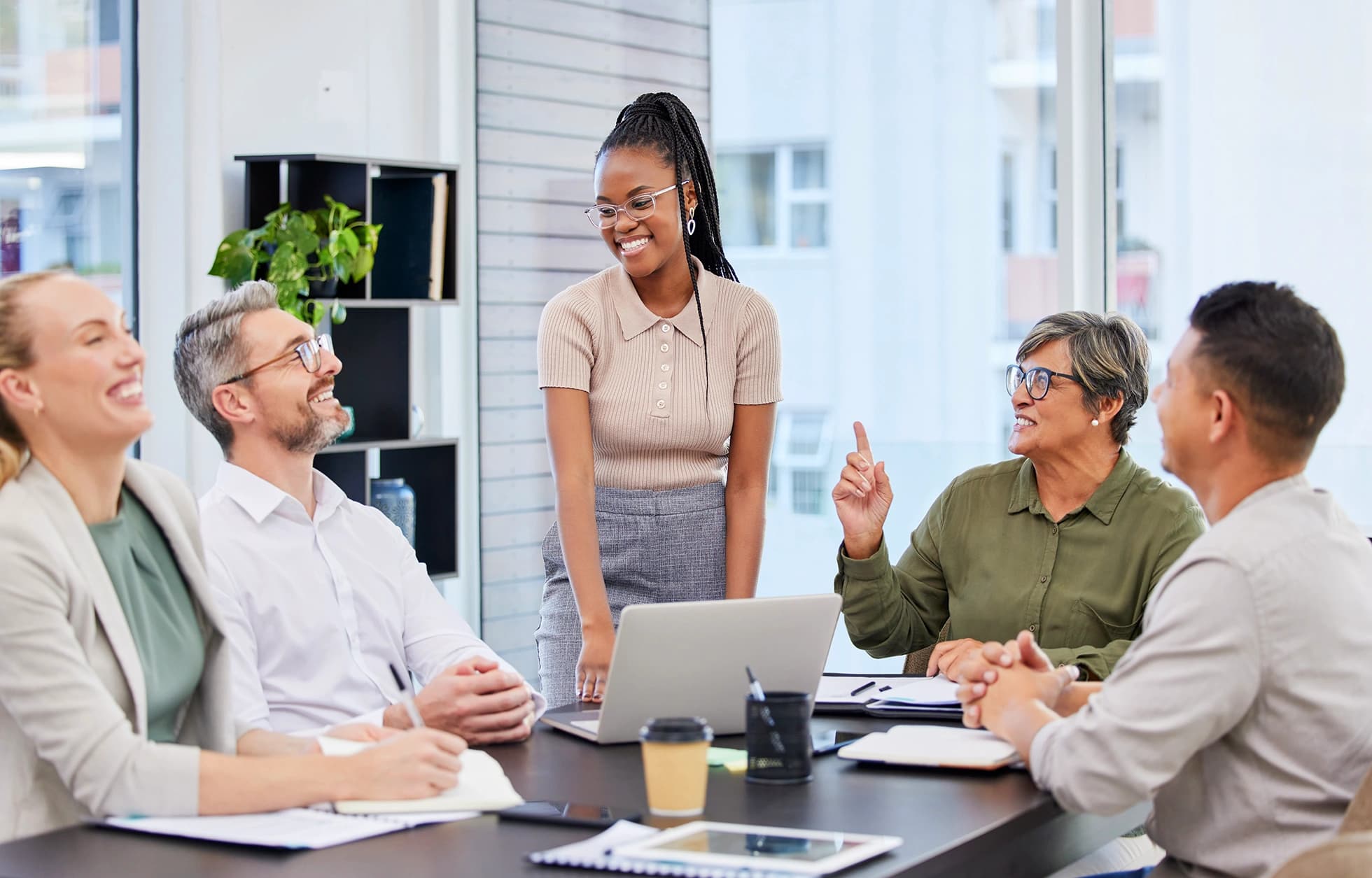 A group of five colleagues smiling and talking during a meeting around a conference table in a bright, modern office.